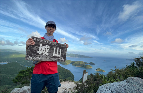 高い山の上で「城山」と書いてある板をもって写真に写る男性
